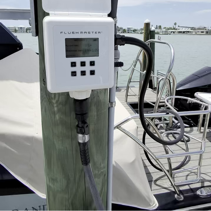 A white Flushmaster water treatment device mounted on a wooden post on a boat, with a screen display and control buttons, set against a background of water and dock.
