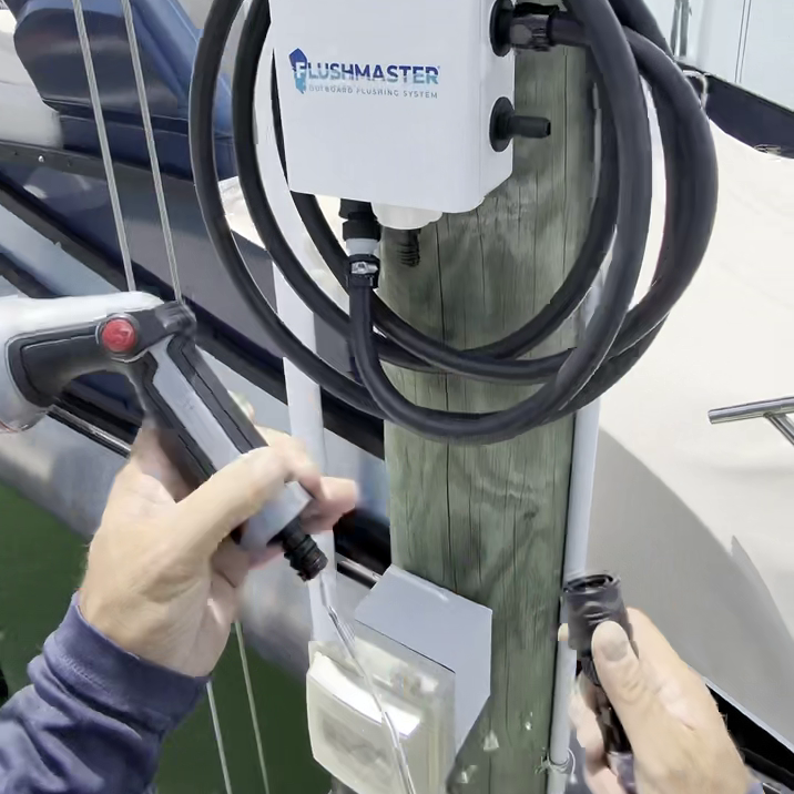 A white Fishmaster marine water system controller mounted on a wooden post, with a coiled black hose and a hose connector being attached by two hands on a boat.
