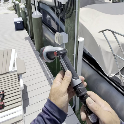A close-up of a person's hands holding a black and gray wand-style pressure washer nozzle at a boat dock, with a covered boat and wooden pilings in the background.