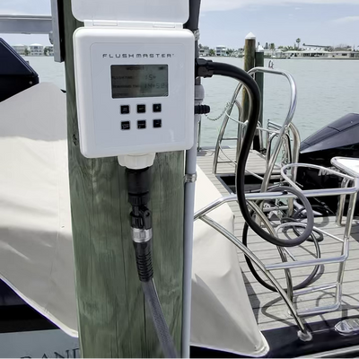 A white Flushmaster water treatment device mounted on a wooden post on a boat, with a screen display and control buttons, set against a background of water and dock.